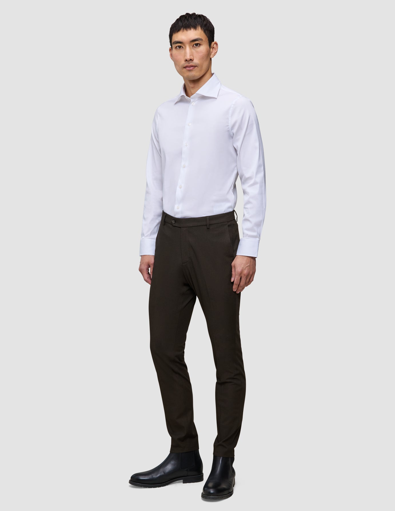 En mann har på seg en hvit skjorte, Motion Suit Pants Slim Cognac og svarte støvler i et rent, skandinavisk design.